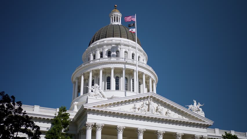 California State Capitol building in Sacramento. The sun shines brightly as flags wave above the dome.