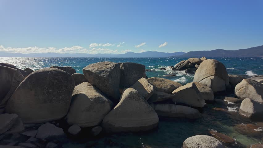Granite rocks on the shore of Lake Tahoe with clear turquoise water and mountain background in California. Scenic natural landscape with waves and blue sky, ideal for travel and outdoor themes