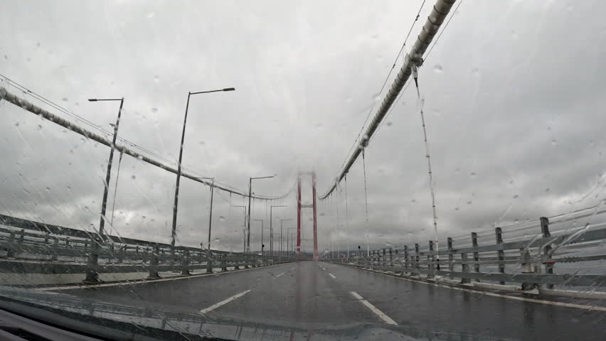 Approaching red suspension bridge on rainy day. Vehicle traversing wet asphalt roadway, approaching large red suspension bridge amid cloudy, rainy conditions, showcasing transportation infrastructure