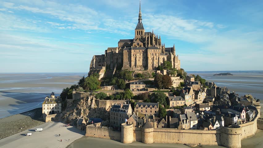 A drone view of Mont-Saint-Michel Abbey nestled in expansive tidal landscape with rural land, on a sunny day with cloudy sky in France