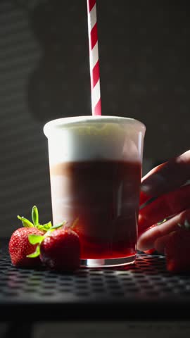 A hand picks up a freshly prepared layered strawberry milk drink with chocolate syrup, milk foam, and a paper straw. Fresh strawberries decorate the scene in a bright café-style setup.