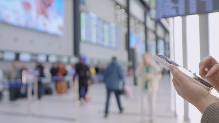 Close-up of hands holding and using a digital tablet. Blurred background shows travelers moving through a busy airport terminal. People walk with luggage, some check phone or talk