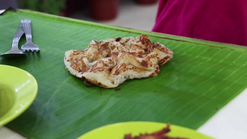 Close-up of person pouring generous amount of curry onto roti canai or prata served on banana leaf at an Indian restaurant in Malaysia.