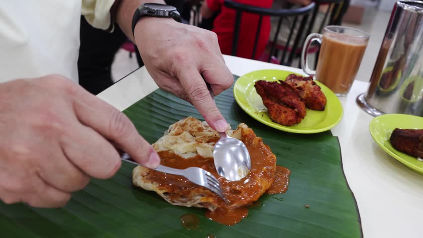 Close-up of person enjoying roti canai with curry gravy and fried chicken served on banana leaf using spoon and fork in an Indian restaurant in Malaysia. Roti canai is popular breakfast among Malaysia