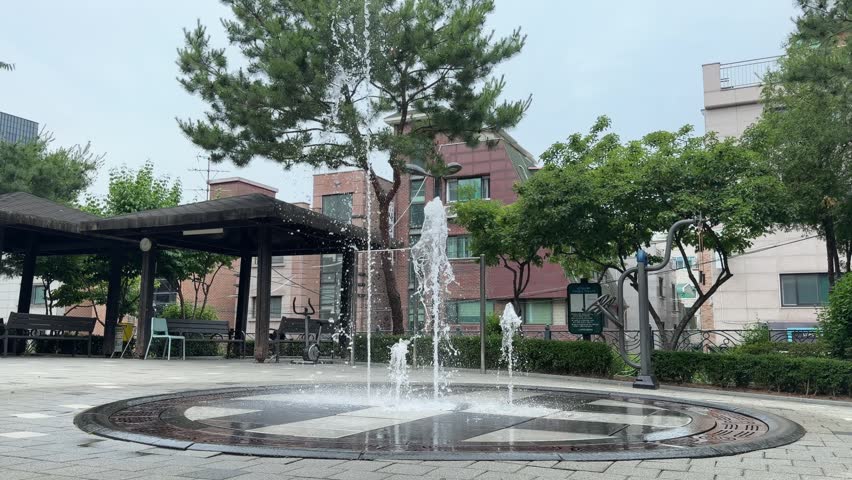 Water fountain spraying upward in a sunny city park surrounded by lush green trees