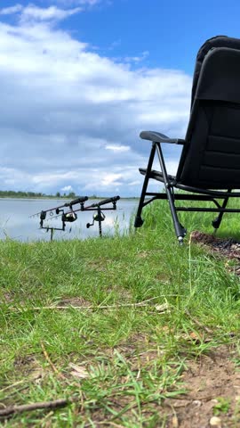 Fishing rods are set up on stands next to a calm lake, with a folding chair and lush grass under a bright blue sky filled with clouds