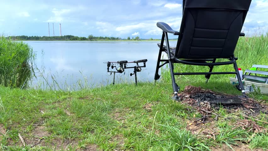 Fishing rods are set on a stand beside a serene lake, with a folding chair and vibrant grass framing the peaceful water scene