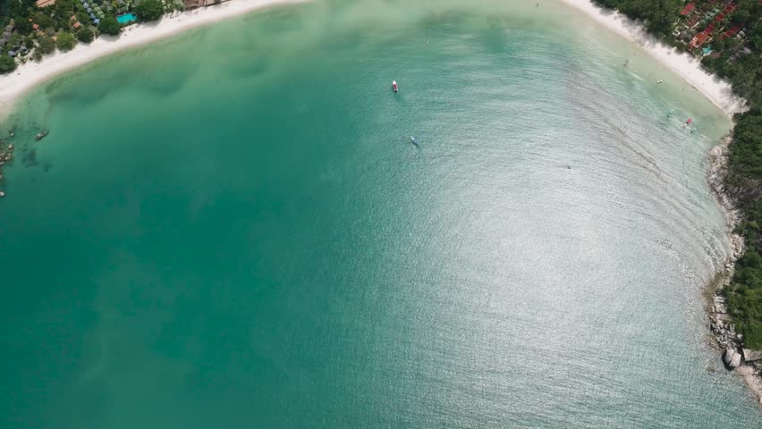 Turquoise water reflecting sunlight near white sand beach with small resorts visible along the coastline. Thailand, Koh Phangan. Thong Nai Pan Yai Beach.