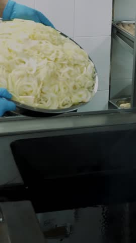 Close-up, a cook in gloves puts a lot of fresh, chopped onion on a large industrial frying pan for roasting, cooking. mixes onions when roasting. process of cooking in mess hall, restaurant.