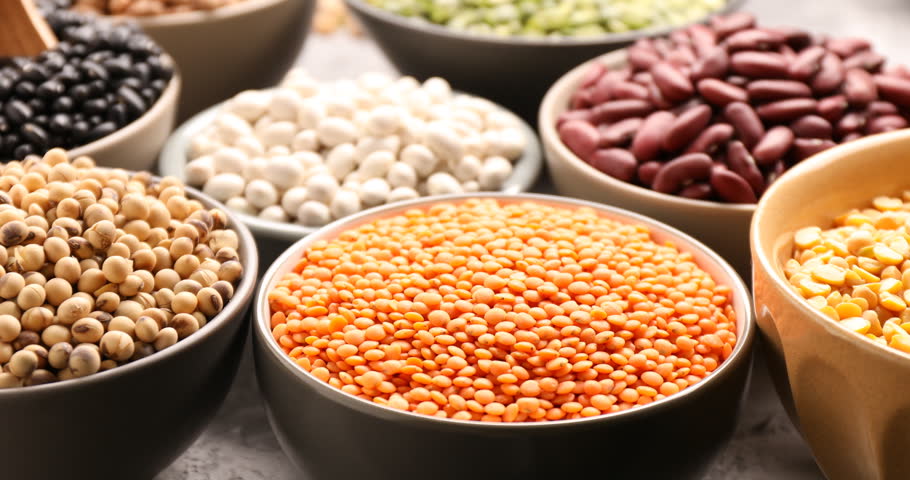 Pouring lentils into bowl from scoop, different types of legumes on grey table, closeup