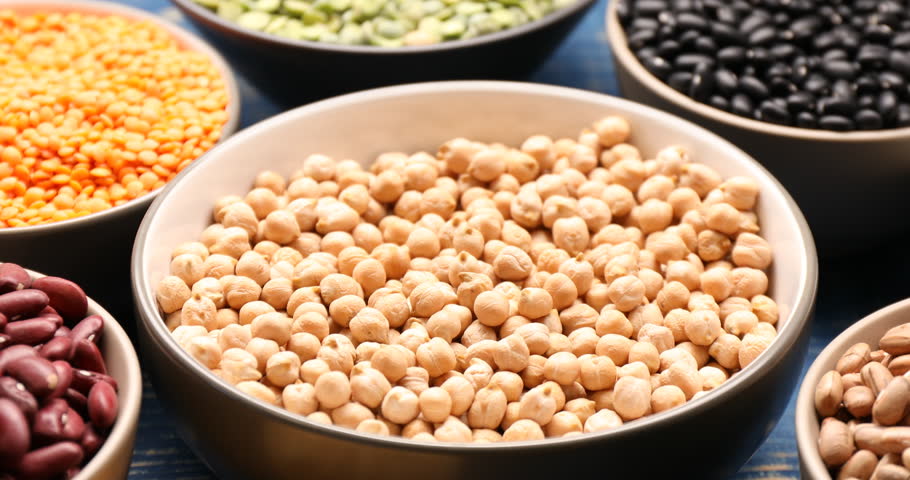 Pouring chickpeas from scoop into bowl, different types of legumes on blue wooden table, closeup