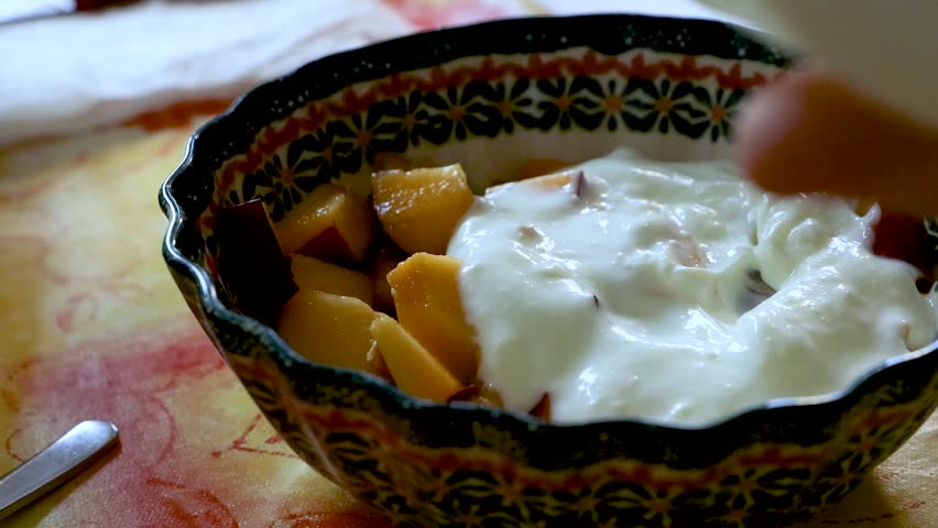Spoon adding yogurt to a bowl of cut peach pieces and stirring them together. 
