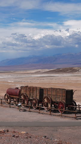 Vertical Screen: Death Valley National Park Borax Factory historic twenty mule team wagons displayed in vast desert landscape with dramatic clouds over distant mountains and salt flats in California.
