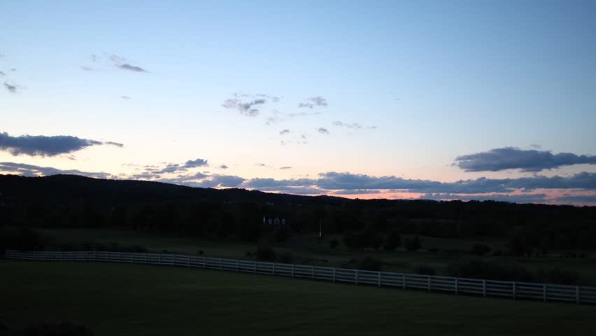 Peaceful rural landscape at dusk with a wide grassy field, white fence, dark hills, and soft clouds in a fading blue and pink evening sky.
