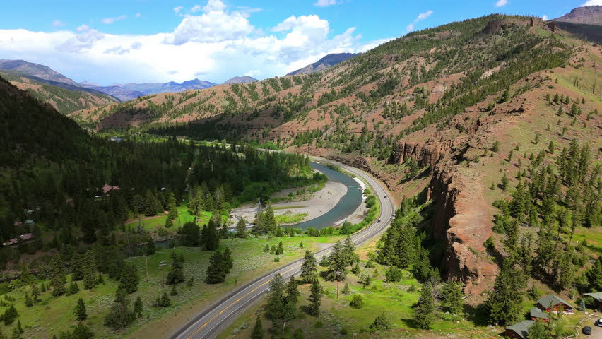 Aerial view of scenic Montana mountain valley featuring a winding river alongside a curved highway, with lush green pine forests, dramatic red rock cliffs, and rolling hills under blue skies.