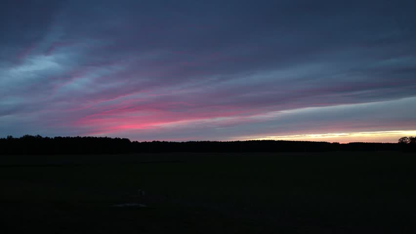 Dark open field under a dramatic sunset sky with pink and purple clouds, a low horizon, and a calm rural landscape at dusk.