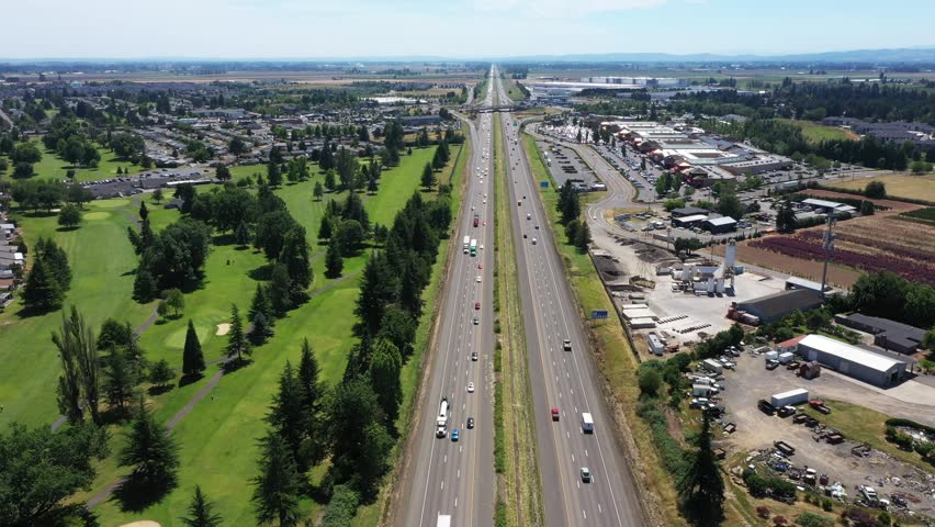 4K aerial drone video footage flying above Interstate 5 near Woodburn, Oregon, USA on a clear summer day. The scene shows steady freeway traffic moving through the Willamette Valley, with green spaces, suburban neighborhoods, commercial buildings, and the Woodburn outlet shopping center visible beside the highway. This wide aerial view can be used for themes related to transportation, travel, infrastructure, commuting, road traffic, retail development, regional growth, commercial districts, highway systems, and summer travel in the Pacific Northwest.