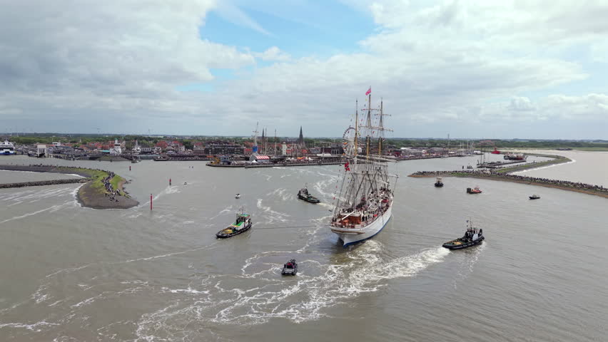 Large traditional tall ship enters Harlingen harbor with tugboats and small boats on the Wadden Sea coast of Friesland, Netherlands.