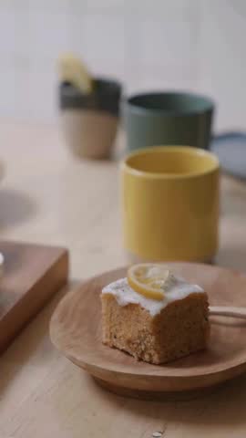 A slice of cake topped with white icing and a lemon wedge sits on a wooden plate alongside a yellow mug.