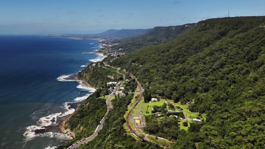 Aerial view of Sea Cliff Bridge winding along the rugged coastline with lush green cliffs and the blue ocean in Clifton, New South Wales, Australia.