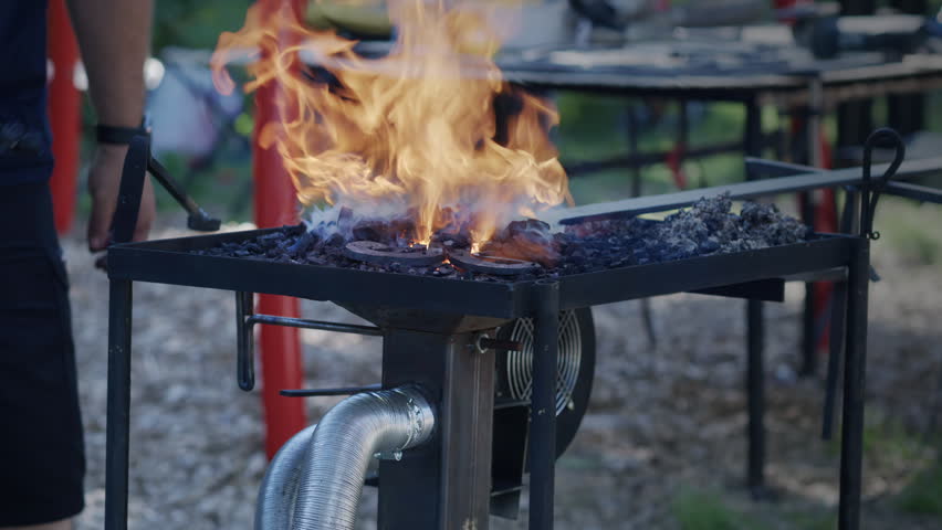 A blacksmith adds coal and water to a coal forge as flames, smoke, and embers rise from the fire bed.
