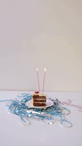 A festive slice of layered cake sits on a white plate with two lit pink candles and colorful party streamers.