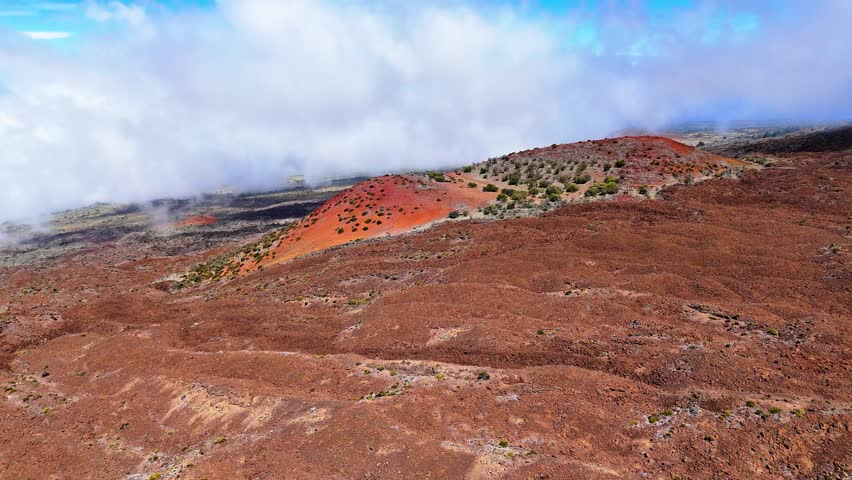 Aerial view of the volcanic Mauna Kea cinder cone with rugged lava fields and scattered vegetation under a cloudy sky in Hawaii County, Hawaii, United States.