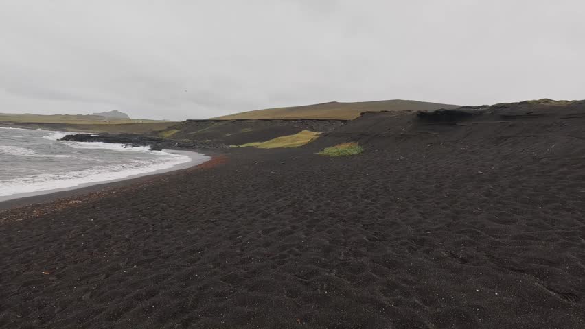 A black sand beach with waves and overcast dramatic sky on a rainy summer day in Vestmannaeyjar Island, Iceland