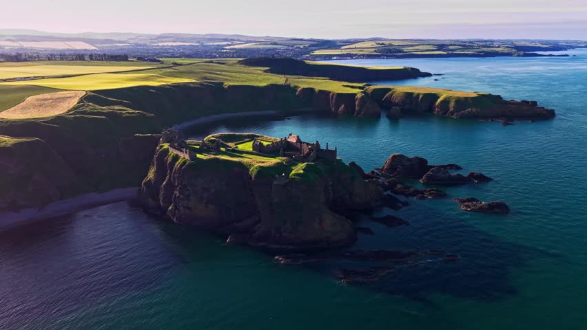 Aerial view of Dunnottar Castle ruins on dramatic sea cliffs at sunset on the Aberdeenshire coast, Scotland, with blue ocean and rugged shoreline. 4K UHD video.