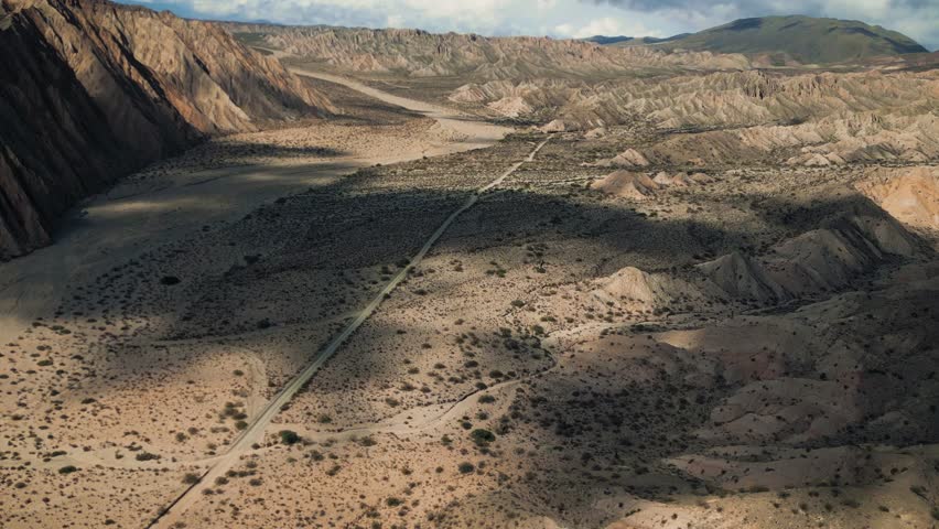 Aerial view of the Valles Calchaquies desert landscape with a winding road cutting through rugged mountains and dramatic shadows in San Carlos, Salta, Argentina.