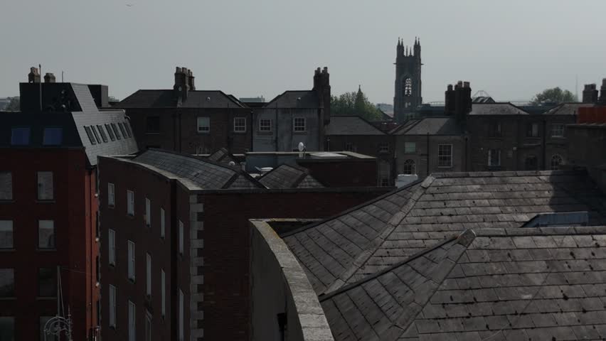 mooth aerial pan across the historic Georgian rooftops of Dublin, Ireland, revealing rows of brick chimney stacks and slate roofs with the Aviva Stadium visible on the skyline. The footage contrasts Dublin