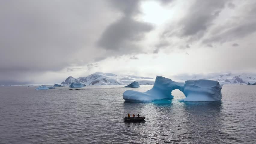 Aerial view of a large iceberg with a natural arch floating in the cold waters of Antarctica, with a small boat carrying people nearby and snowy mountains in the distance.