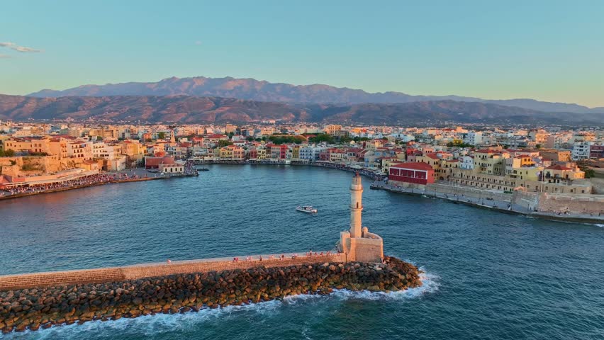 Aerial view of the historic Old Venetian Port of Chania with its iconic lighthouse, colorful waterfront buildings, and mountains in the background in Chania, Greece.