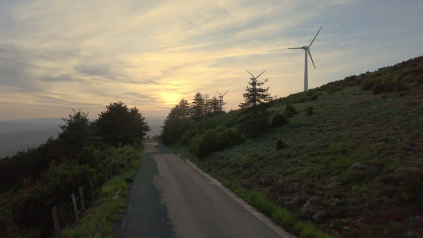A peaceful countryside path curves with grassy slopes hills and windmills at sunset