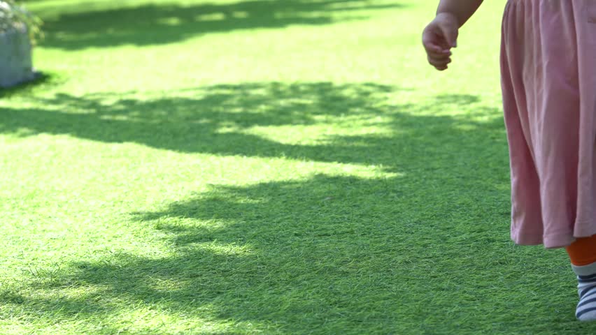 A close up view of a child walking across a vibrant green grass lawn in a sunlit park with soft tree shadows.