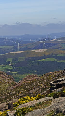 Wind turbines stand on rolling hills in a rural mountain landscape. Rocky foreground and patchwork fields frame the wind farm under a cloudy sky.