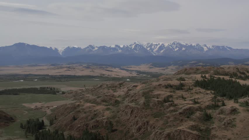 Wide aerial landscape of the Kurai Steppe with dry hills, river channels and distant snowcapped Altai Mountains under cloudy sky, Altai Republic, Russia