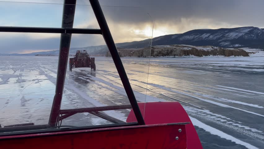 Point of view from a vehicle driving on the icy surface of Lake Baikal in Siberia, Russia, with snow tracks, rocky shoreline and distant mountains under a winter sunset