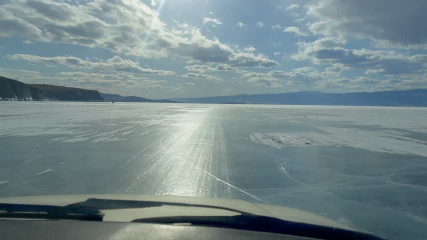 POV view from a vehicle driving on the frozen surface of Lake Baikal with sun glare, textured ice and distant mountains under a cloudy sky in Russia