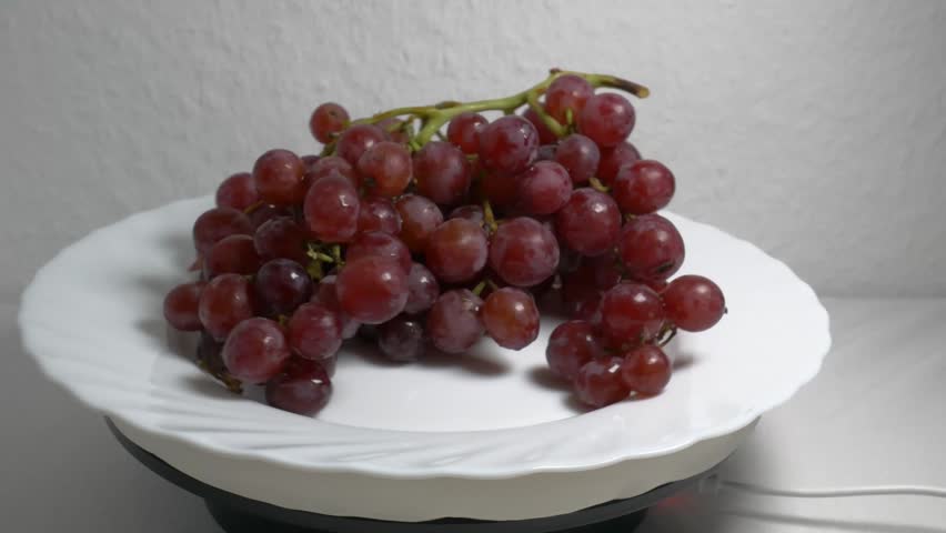 Close-up of a fresh bunch of red grapes rotating on a white turntable, showcasing their ripe berries and glossy texture under studio lighting.