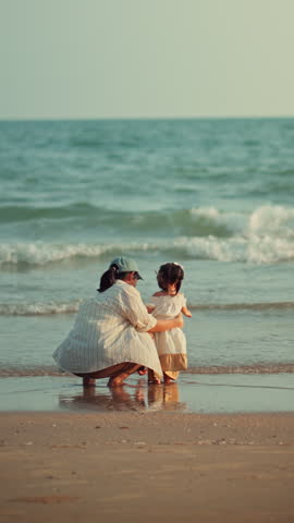 Back view of Asian mother crouching and hugging her little daughter on sandy beach, happy family looking at sea waves during summer vacation for travel tourism, parenting, and childhood lifestyle concept