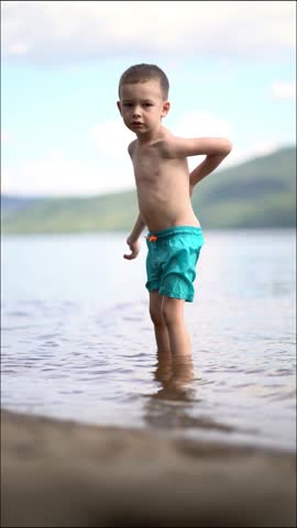 A young boy joyfully kicks and splashes water with his bare foot at the sunny lake shore. He smiles with arms wide open while playing on a summer day.