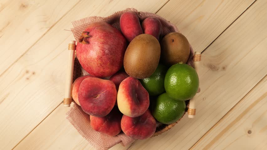 Wicker basket full of ripe peaches, pomegranates, kiwis, and limes spinning on a wooden surface