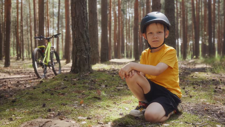 Full body shot of an eight year old boy in a bike helmet sitting on moss in a sunny pine forest and showing thumbs up with his bicycle near a tree behind him. High quality 4k footage