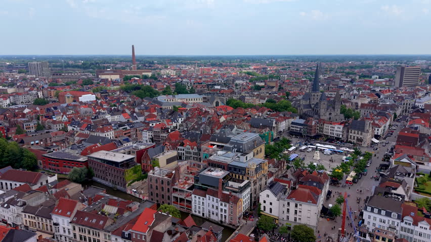 A magnificent wide-angle aerial photograph capturing the intricate urban fabric of a historic European city (Tournai). The foreground and midground are densely packed with charming traditional old town buildings featuring distinctive red-tiled roofs, brick facades, and narrow winding streets. On the right, a large public city square or market plaza opens up, filled with market stalls, white tents, outdoor cafes, and tiny figures of people walking. Rising majestically above the square is a grand historic stone cathedral and church complex with prominent medieval towers and a tall, dark spire piercing the sky. In the far background, a large industrial brick chimney stands tall, while the urban landscape and residential neighborhoods stretch out toward a flat horizon under a bright, softly overcast sky, beautifully combining historical architecture and vibrant city life.