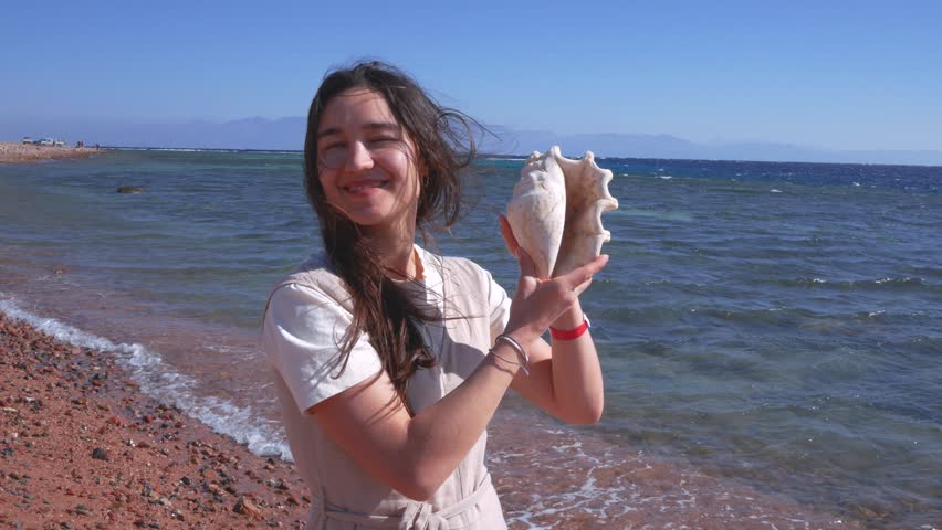 Happy young woman listening to the sound of the ocean in a seashell on a sunny beach