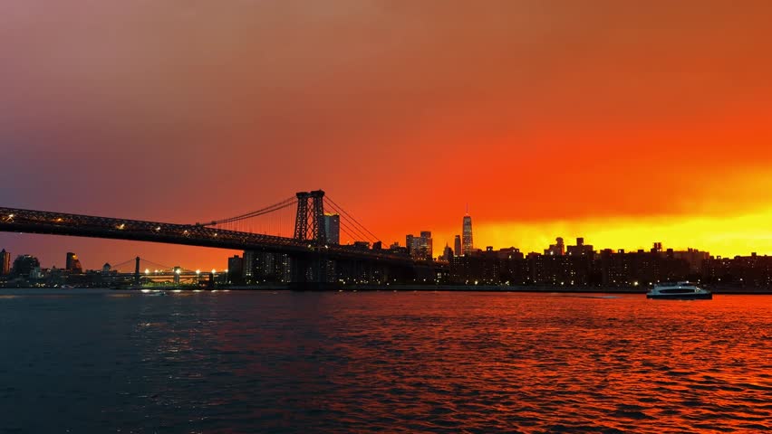 Williamsburg Bridge beside Lower Manhattan skyline at dusk. Bridge structure crosses left frame, with red sky, rippled water, and city lights.
