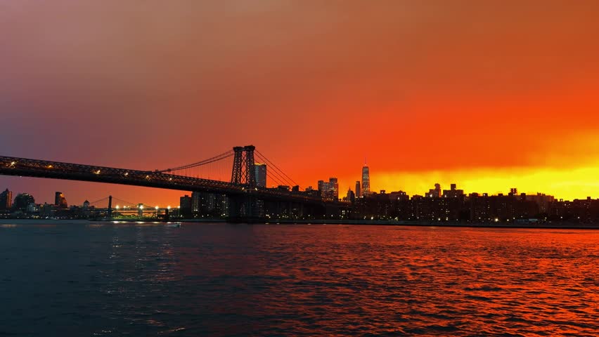 Williamsburg Bridge and Manhattan waterfront under evening clouds. Wide river scene with steel truss bridge, lit buildings, and yellow orange horizon.