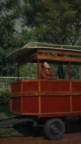 Red wooden cart with curved roof and open sides. Person sits calmly, looking toward the background. Green trees and fence create natural setting
