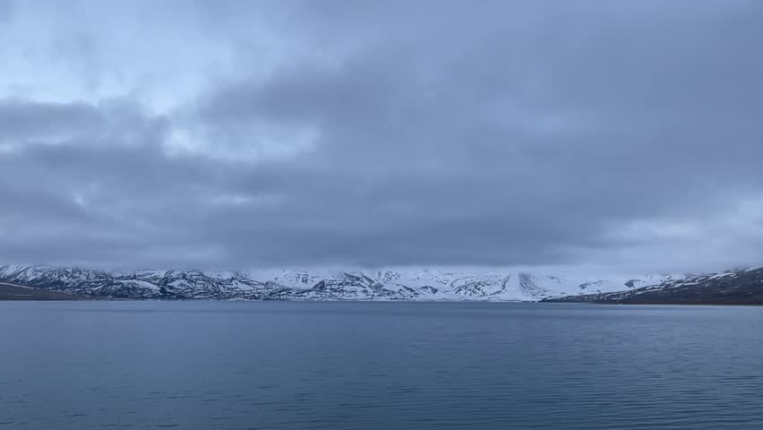A tranquil mountain lake reflects the dramatic, snow-capped peaks under a perpetually cloudy sky.